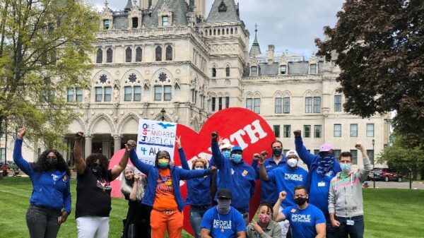ACLUCT Smart Justice leaders, in blue people not prisons shirts, crouch and stand with a massive red heart that says "strength" in white letters. Behind them is the CT capitol building. They are masked and raising their fists in celebration and power.