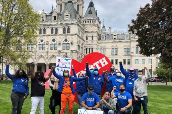 ACLUCT Smart Justice leaders, in blue people not prisons shirts, crouch and stand with a massive red heart that says "strength" in white letters. Behind them is the CT capitol building. They are masked and raising their fists in celebration and power.