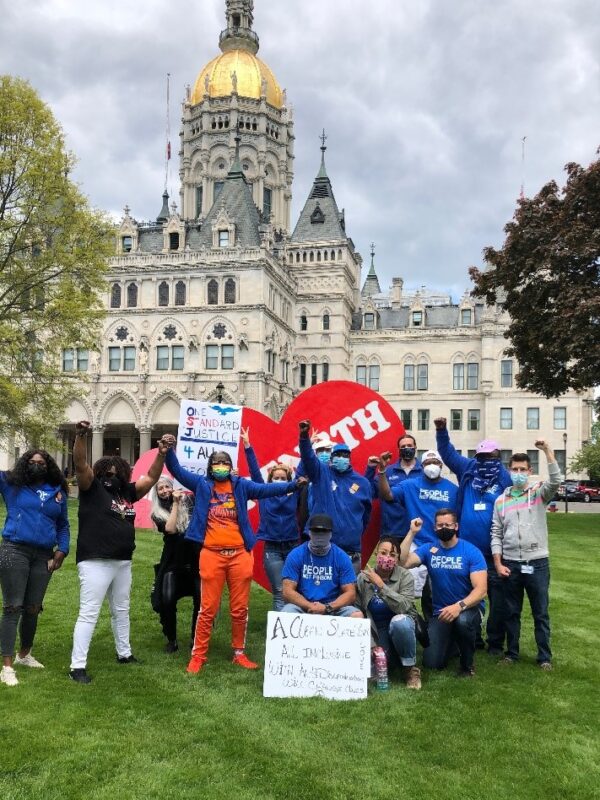 ACLUCT Smart Justice leaders, in blue people not prisons shirts, crouch and stand with a massive red heart that says "strength" in white letters. Behind them is the CT capitol building. They are masked and raising their fists in celebration and power.