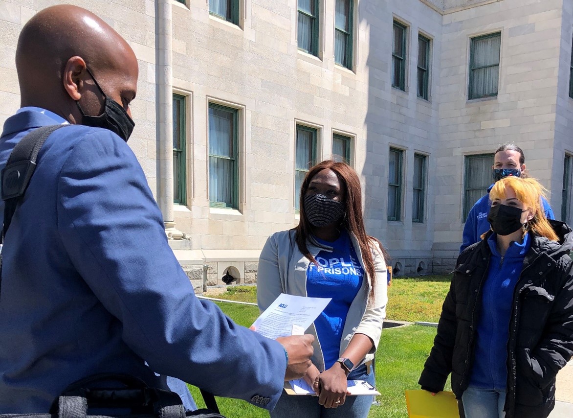 ACLUCT smart justice leaders Shelby and Marquita hand a legislator a copy of a Smart Justice factsheet. In the foreground, the legislator hold the paper, wearing a mask, glancing down to read. In the background and center, Shelby and Marquita