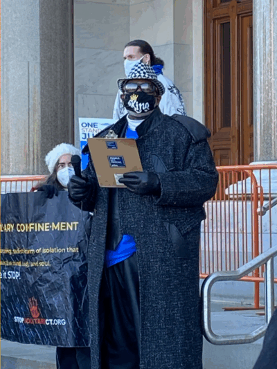 ACLUCT smart justice leader Curtis B. Hudson stands in front of the CT Capitol steps. He is wearing a black and white fedora, sunglasses, black mask, black trenchcoat, and blue Smart Justice shirt, and black gloves, and he is holding a clipboard.