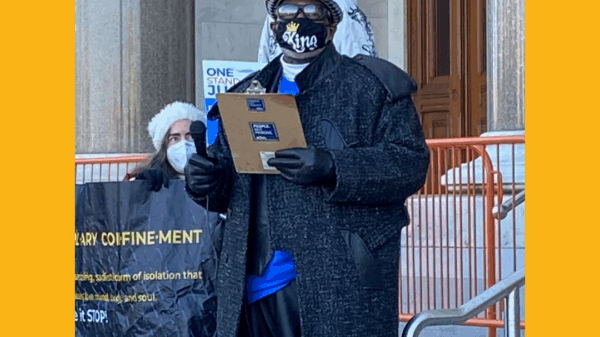 ACLUCT smart justice leader Curtis B. Hudson stands in front of the CT Capitol steps. He is wearing a black and white fedora, sunglasses, black mask, black trenchcoat, and blue Smart Justice shirt, and black gloves, and he is holding a clipboard.