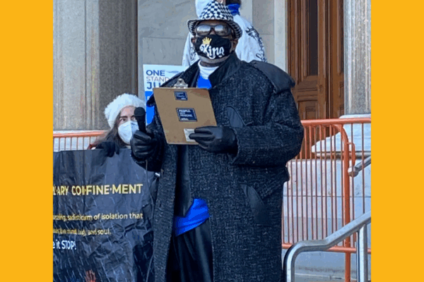 ACLUCT smart justice leader Curtis B. Hudson stands in front of the CT Capitol steps. He is wearing a black and white fedora, sunglasses, black mask, black trenchcoat, and blue Smart Justice shirt, and black gloves, and he is holding a clipboard.