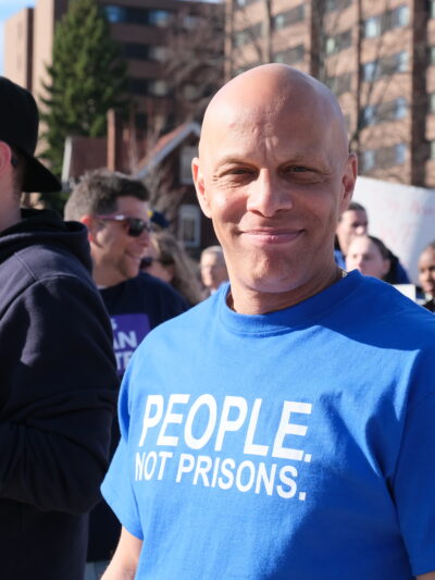 ACLUCT smart justice leader Brian Sullivan smiles at the camera. He's wearing a blue people not prisons shirt. Behind him is a crowd of people at a protest in Feb. 2020.