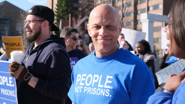 ACLUCT smart justice leader Brian Sullivan smiles at the camera. He's wearing a blue people not prisons shirt. Behind him is a crowd of people at a protest in Feb. 2020.