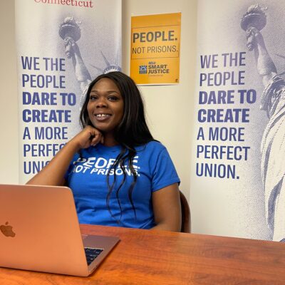 ACLUCT Smart Justice leader Shelby Henderson sits behind a desk, smiling at the camera, with one hand on her chin. She is behind a desk, laptop and wearing a blue people not prisons shirt, with ACLU of CT banners and a people not prisons poster behind her