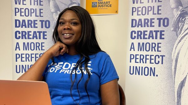 ACLUCT Smart Justice leader Shelby Henderson sits behind a desk, smiling at the camera, with one hand on her chin. She is behind a desk, laptop and wearing a blue people not prisons shirt, with ACLU of CT banners and a people not prisons poster behind her