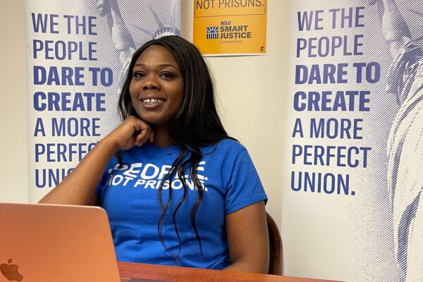 ACLUCT Smart Justice leader Shelby Henderson sits behind a desk, smiling at the camera, with one hand on her chin. She is behind a desk, laptop and wearing a blue people not prisons shirt, with ACLU of CT banners and a people not prisons poster behind her