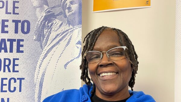 ACLUCT smart justice leader Terri Ricks sits, smiling and looking straight at the camera. She is wearing a blue ACLU of Connecticut Smart justice hoodie and glasses. Behind her is a yellow people not prisons poster.