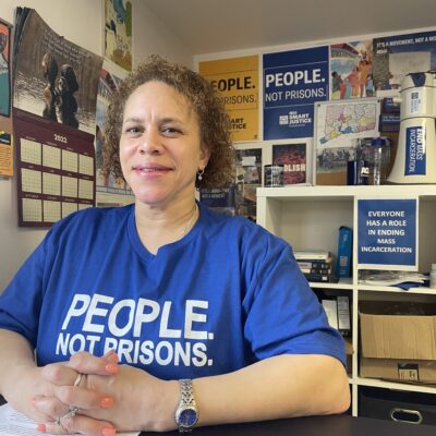 ACLU of Connecticut Smart Justice leader Pierlette Jones sits behind a desk, hands folded, smiling at the camera. She is wearing a blue people not prisons shirt and a watch. A copy of legislative testimony is under her clasped hands.