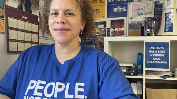 ACLU of Connecticut Smart Justice leader Pierlette Jones sits behind a desk, hands folded, smiling at the camera. She is wearing a blue people not prisons shirt and a watch. A copy of legislative testimony is under her clasped hands.