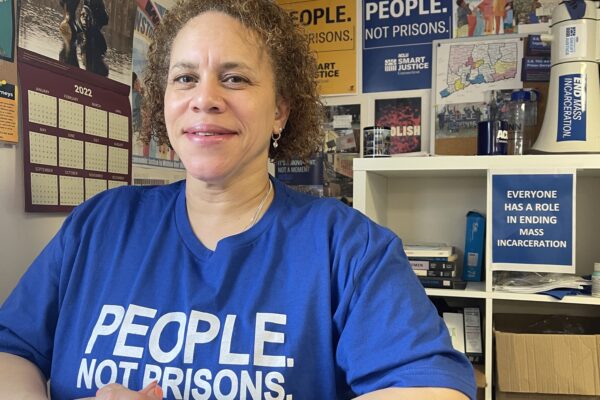 ACLU of Connecticut Smart Justice leader Pierlette Jones sits behind a desk, hands folded, smiling at the camera. She is wearing a blue people not prisons shirt and a watch. A copy of legislative testimony is under her clasped hands.