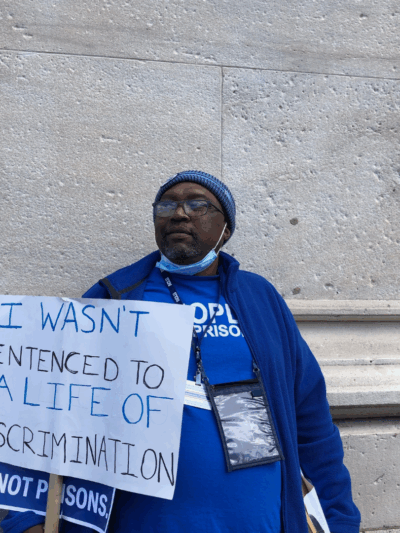 ACLUCT senior policy organizer Anderson Curtis stands with a white sign that says "I wasn't sentenced to a lifetime of discrimination." He wears a blue people not prisons shirt and sweatshirt and blue hat, glasses. He is leaning on a gray stone building
