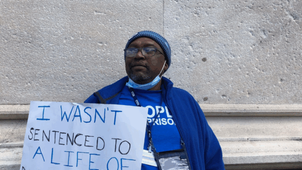ACLUCT senior policy organizer Anderson Curtis stands with a white sign that says "I wasn't sentenced to a lifetime of discrimination." He wears a blue people not prisons shirt and sweatshirt and blue hat, glasses. He is leaning on a gray stone building