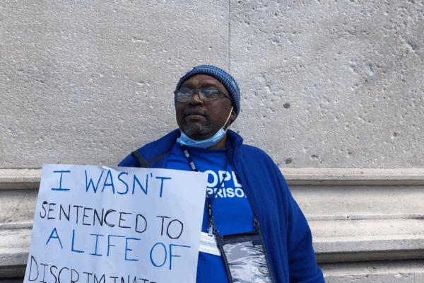 ACLUCT senior policy organizer Anderson Curtis stands with a white sign that says "I wasn't sentenced to a lifetime of discrimination." He wears a blue people not prisons shirt and sweatshirt and blue hat, glasses. He is leaning on a gray stone building