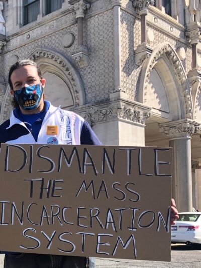 ACLUCT smart justice leader Will stands with a cardboard sign that says, in black and white, "dismantle the mass incarceration system." Behind him is the CT Capitol. he is wearing a mask and a sweatshirt, and yellow people not prison pin