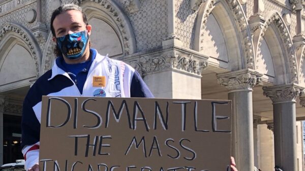 ACLUCT smart justice leader Will stands with a cardboard sign that says, in black and white, "dismantle the mass incarceration system." Behind him is the CT Capitol. he is wearing a mask and a sweatshirt, and yellow people not prison pin