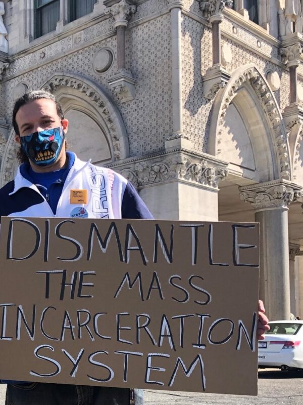 ACLUCT smart justice leader Will stands with a cardboard sign that says, in black and white, "dismantle the mass incarceration system." Behind him is the CT Capitol. he is wearing a mask and a sweatshirt, and yellow people not prison pin