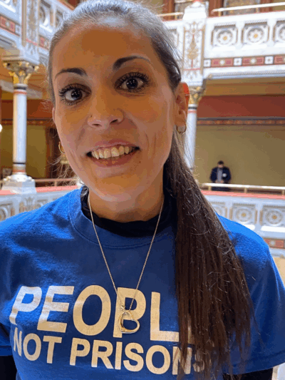 Image description: Alex Brown, ACLU-CT Smart Justice leader, stands, smiling directly at the camera. She is wearing a blue People Not Prisons t-shirt and black long-sleeved shirt under it. Her long brown hair is in a ponytail.