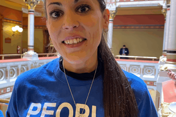 Image description: Alex Brown, ACLU-CT Smart Justice leader, stands, smiling directly at the camera. She is wearing a blue People Not Prisons t-shirt and black long-sleeved shirt under it. Her long brown hair is in a ponytail.