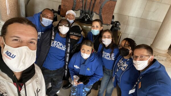 ACLU-CT Smart Justice leaders crouch in front of the bell at the CT State Capitol. They are wearing masks and blue people not prisons shirts, and smiling at the camera.