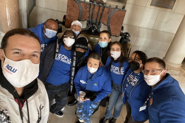 ACLU-CT Smart Justice leaders crouch in front of the bell at the CT State Capitol. They are wearing masks and blue people not prisons shirts, and smiling at the camera.