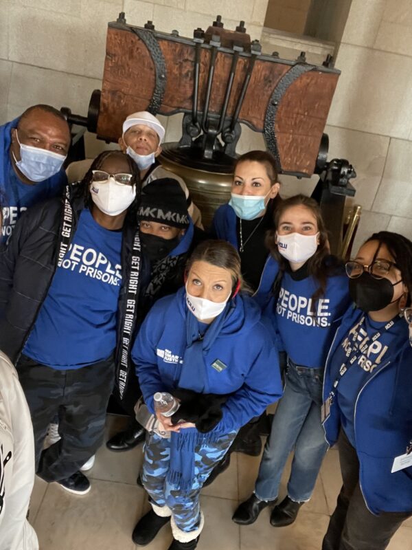 ACLU-CT Smart Justice leaders crouch in front of the bell at the CT State Capitol. They are wearing masks and blue people not prisons shirts, and smiling at the camera.