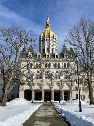 The Connecticut capitol building is centered. The capitol lawn is covered in snow, and a sidewalk leading to the building is clear of snow. The sky is blue and has light clouds.