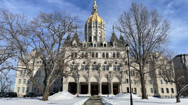 The Connecticut capitol building is centered. The capitol lawn is covered in snow, and a sidewalk leading to the building is clear of snow. The sky is blue and has light clouds.