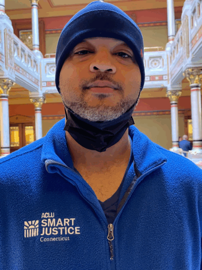 Image description: Tyran Sampson, ACLU-CT Smart Justice leader, stands, facing the camera. He is wearing a blue ACLU of Connecticut Smart Justice zip-up sweatshirt, a navy blue hat, and has a mask pulled down. He looks serious.