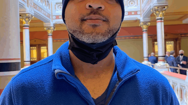 Image description: Tyran Sampson, ACLU-CT Smart Justice leader, stands, facing the camera. He is wearing a blue ACLU of Connecticut Smart Justice zip-up sweatshirt, a navy blue hat, and has a mask pulled down. He looks serious.