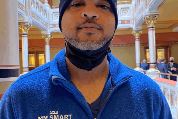 Image description: Tyran Sampson, ACLU-CT Smart Justice leader, stands, facing the camera. He is wearing a blue ACLU of Connecticut Smart Justice zip-up sweatshirt, a navy blue hat, and has a mask pulled down. He looks serious.