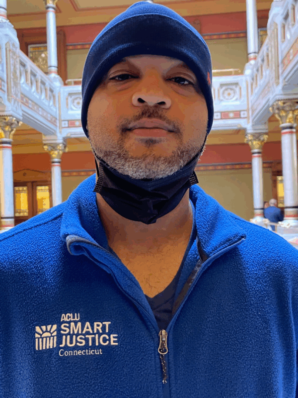 Image description: Tyran Sampson, ACLU-CT Smart Justice leader, stands, facing the camera. He is wearing a blue ACLU of Connecticut Smart Justice zip-up sweatshirt, a navy blue hat, and has a mask pulled down. He looks serious.