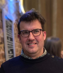 Thomas Regan-Lefebvre looks slightly to the right of the camera. He has brown hair and is wearing glasses, smiling slightly, and wearing a dark blue shirt with a collared shirt underneath.