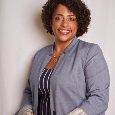 A Black women wearing a grey jacket and vertical striped shirt in front of a white background. The woman is smiling in the image.