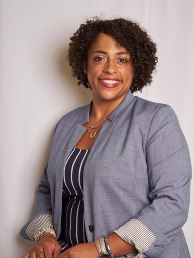 A Black women wearing a grey jacket and vertical striped shirt in front of a white background. The woman is smiling in the image.