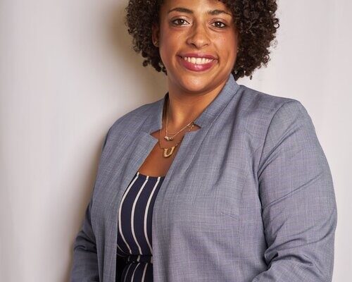 A Black women wearing a grey jacket and vertical striped shirt in front of a white background. The woman is smiling in the image.