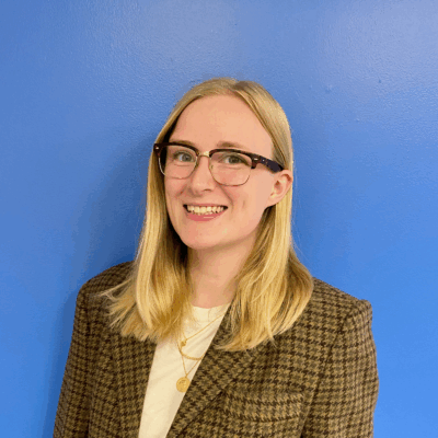 A white woman with blonde hair and glasses smiling at the camera. There is a blue background behind her.