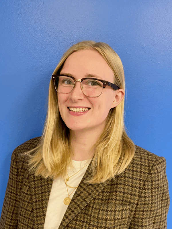 A white woman with blonde hair and glasses smiling at the camera. There is a blue background behind her.