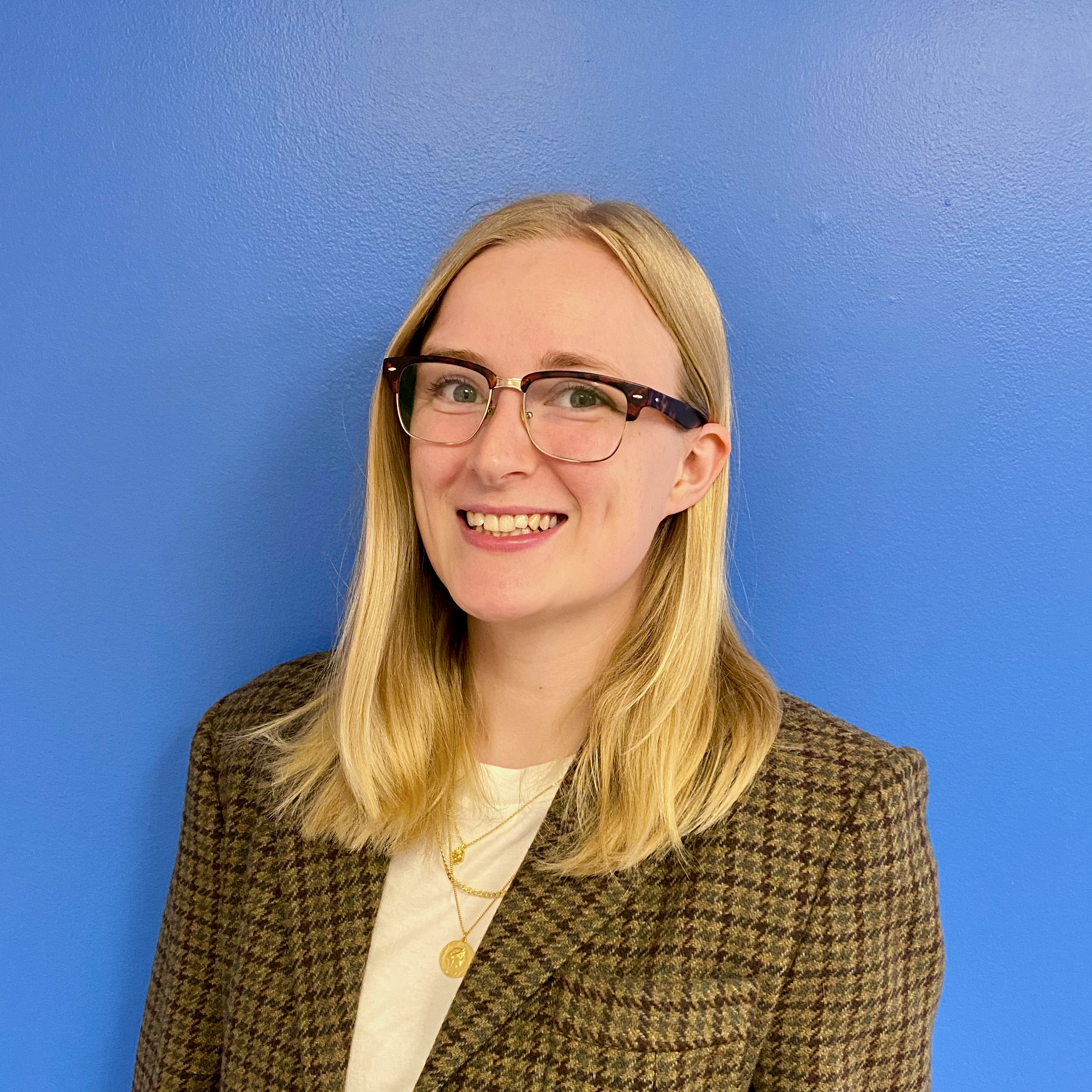 A white woman with blonde hair and glasses smiling at the camera. There is a blue background behind her.