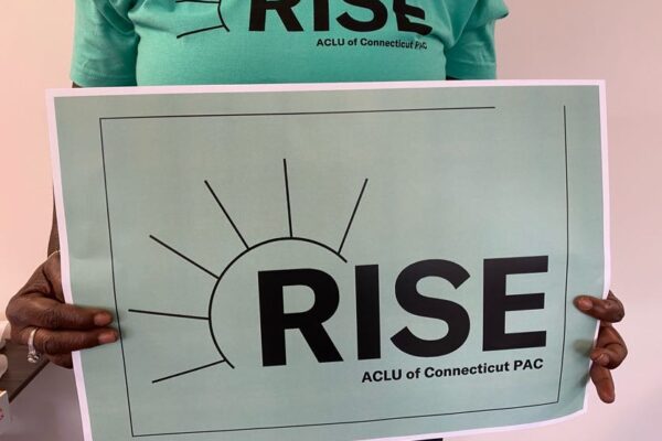A person with long nails and a ring holds an ACLU-CT Rise PAC sign and wears an ACLU-CT Rise PAC shirt. The shirt and sign are statute of liberty green, the ACLUCT Rise PAC logos in black