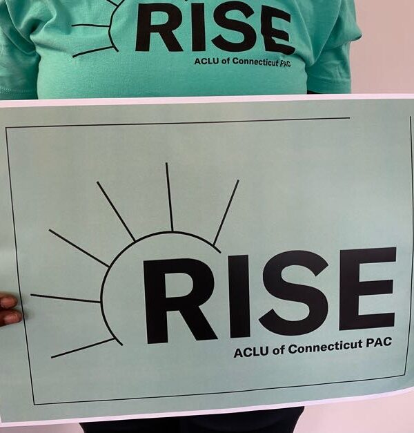 A person with long nails and a ring holds an ACLU-CT Rise PAC sign and wears an ACLU-CT Rise PAC shirt. The shirt and sign are statute of liberty green, the ACLUCT Rise PAC logos in black