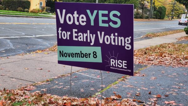 A purple yard sign, with green and white text, says, "vote YES for early voting November 8." the ACLU-CT Rise PAC logo is at the bottom. behind are a street and sidewalk, with autumn leaves scattered around