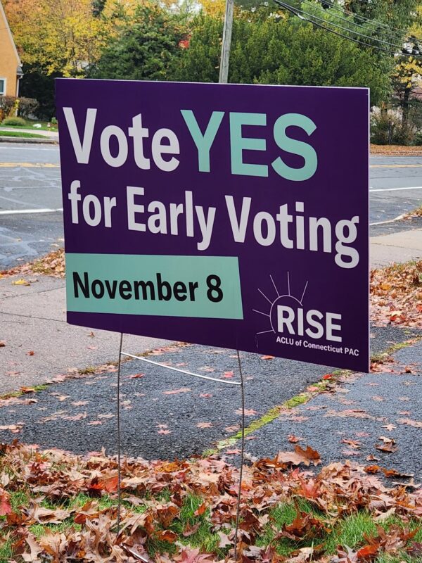 A purple yard sign, with green and white text, says, "vote YES for early voting November 8." the ACLU-CT Rise PAC logo is at the bottom. behind are a street and sidewalk, with autumn leaves scattered around