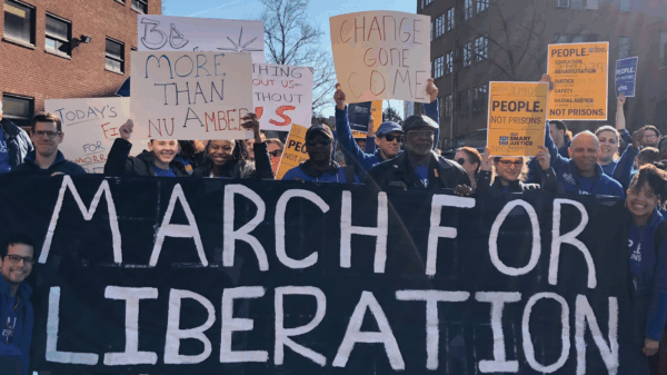 A group of people stand behind a large blue banner with white hand-painted letters that says, "march for liberation." people are holding "people not prisons" signs, handmade signs, and smiling