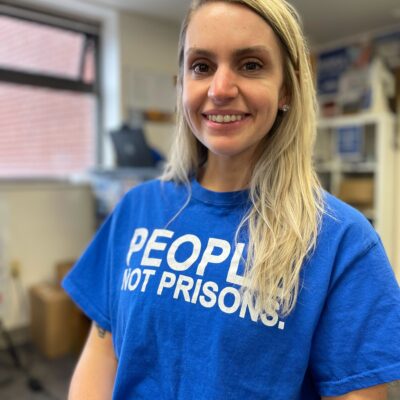 ACLUCT smart justice leader Brittany Lamar stands, smiling and facing the camera. She is wearing a blue people not prisons t-shirt and has long blond hair.