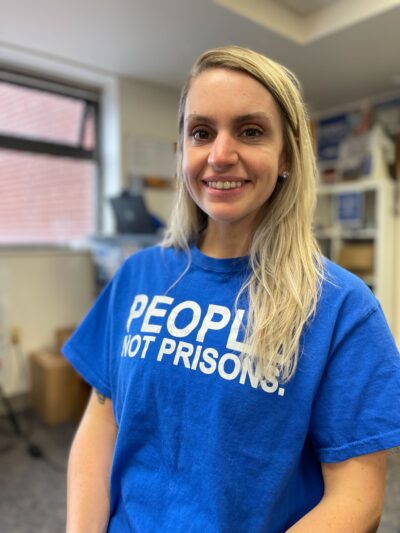 ACLUCT smart justice leader Brittany Lamar stands, smiling and facing the camera. She is wearing a blue people not prisons t-shirt and has long blond hair.