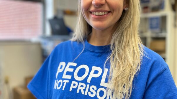 ACLUCT smart justice leader Brittany Lamar stands, smiling and facing the camera. She is wearing a blue people not prisons t-shirt and has long blond hair.