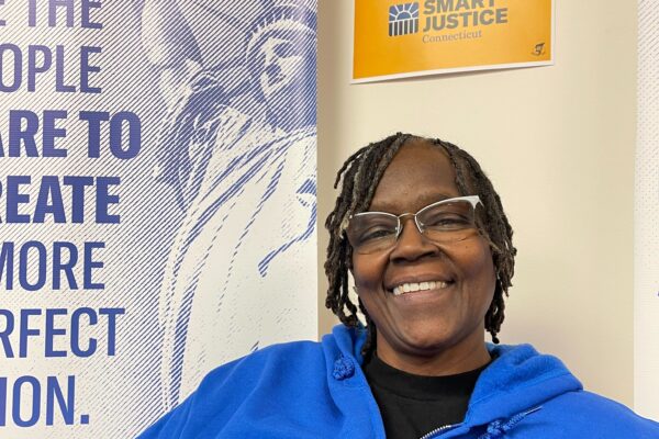 ACLU of CT Smart Justice leader Terri Ricks sits, smiling and facing the camera. She is wearing glasses and a blue people not prisons sweatshirt, in front of a yellow people not prisons sign.