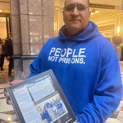ACLU of CT Smart Justice leader Manuel Sandoval faces the camera, standing and holding a framed collage commemorating his LCSW. he is wearing glasses and a blue people not prisons hoodie